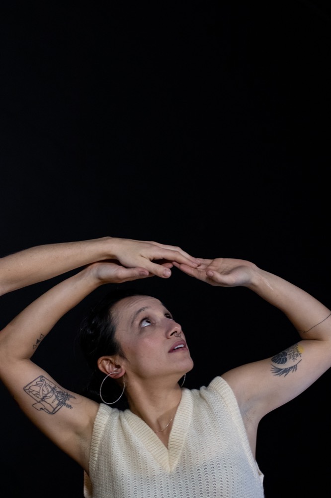 Ayumi Hanada sobre fundo preto, com os braços erguidos acima da cabeça formando um arco e as mãos se tocando, o olhar voltado para cima.