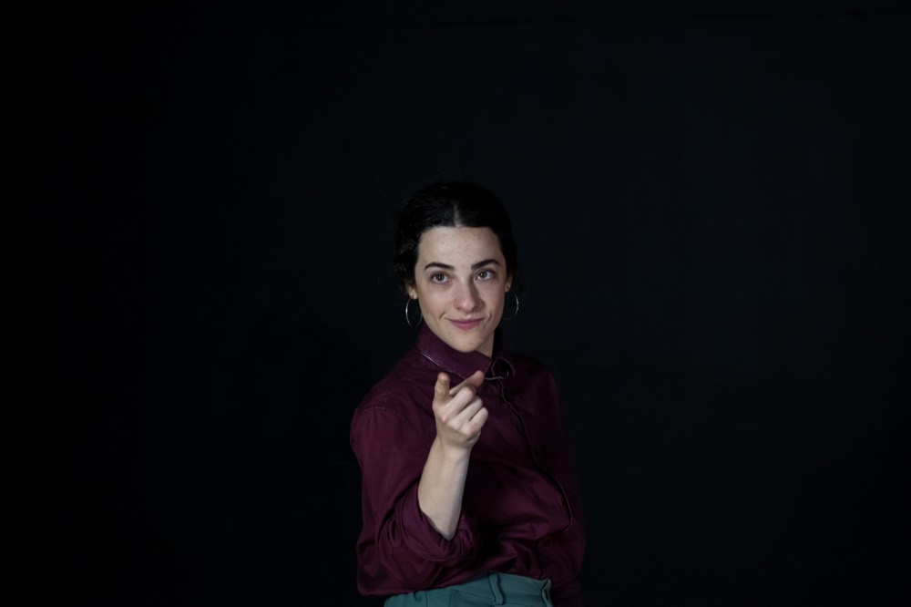 Sabrina Barros sobre fundo preto, de camisa vinho e cabelo preso, apontando o dedo indicador para a câmera.